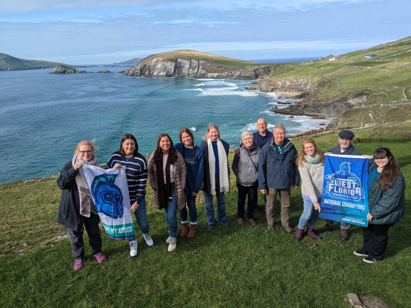 A group of alumni and guests pose in Ireland on the bluffs with a UWF Flag