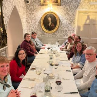 A group of alumni sit inside a dining room with old paintings in the background.