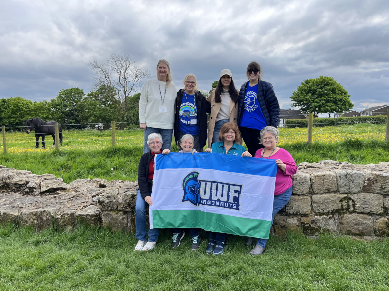Eight travelers pose in a grassy field with a UWF flag.
