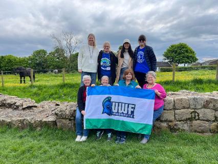 Eight travelers pose in a grassy field with a UWF flag.
