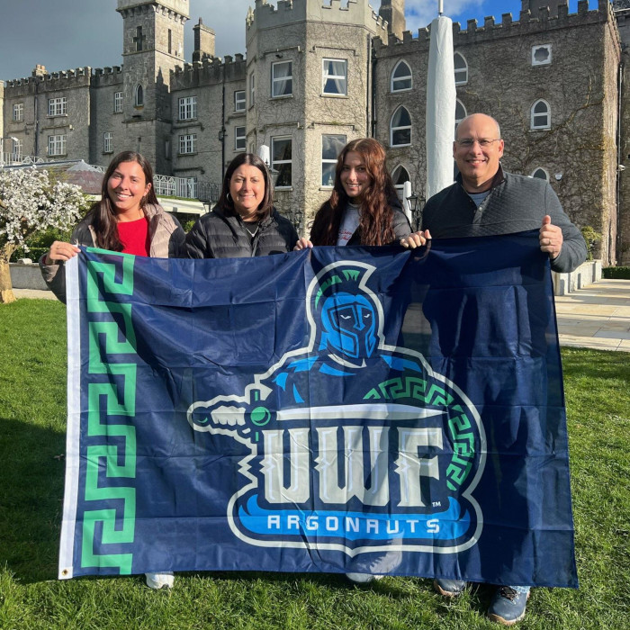 Four alumni and family members pose with a UWF flag in front of a stone castle.