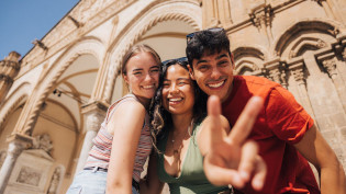 Three young adult travelers flash a peace sign at the camera in Greece.