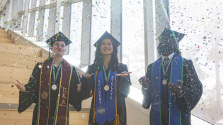 Three graduates in regalia stand in the sun and throw confetti in the air to celebrate.