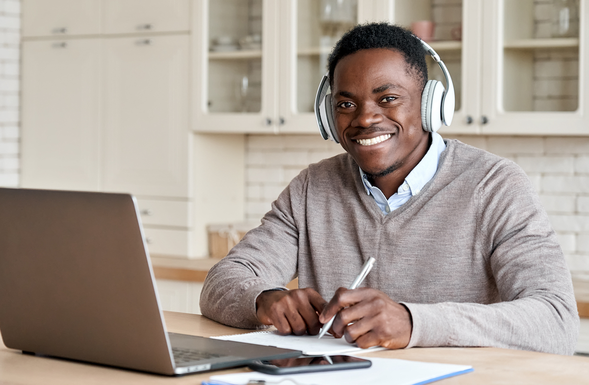 Adult working on online coursework. Smiling and wearing headphones, working on a laptop and taking notes.