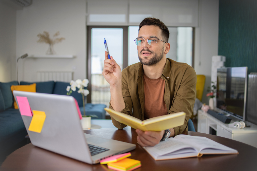Male student holding a book and looking at a laptop