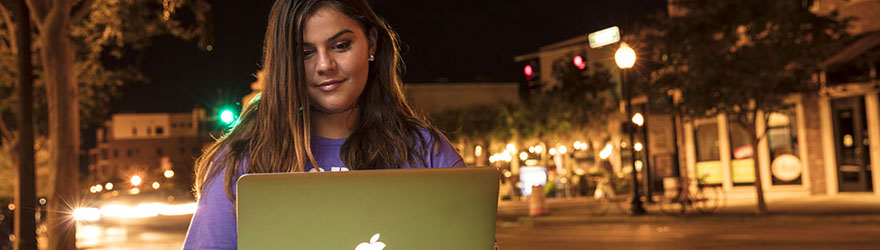 A UWF student smiles while using a laptop in Downtown Pensacola at night.
