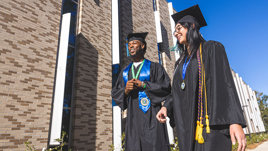 Two students stand in graduation regalia in front of an academic building.