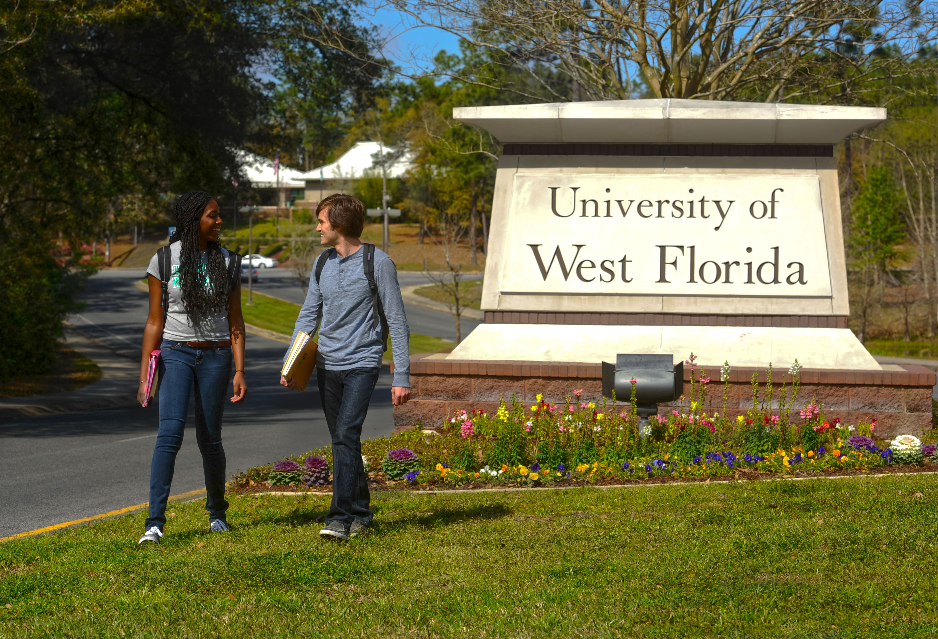 Two students walking by the University of West Florida concrete campus entrance monument display with planted flowers in front of the display.