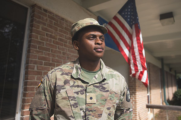 A military student in uniform standing outdoors in front of a brick building that has a United States flag hanging from a pole in the wall.
