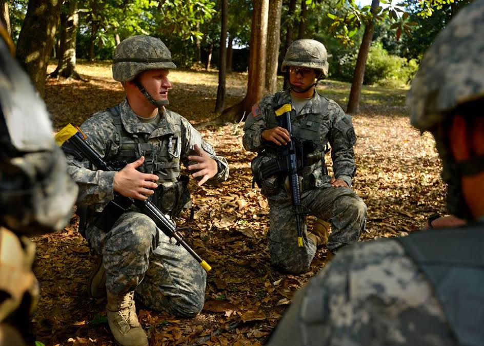 Four military students in military uniform attire holding prop firearms outdoors during a course instruction.