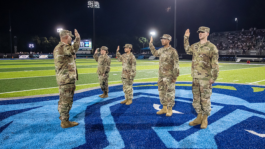 Four military students in military uniform attire raise their right hands in salute during a nighttime oath-taking ceremony on a football field led by a military professor in military uniform attire.