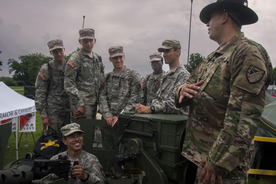 A military faculty in military uniform attire leading an outdoor instruction with a group of six military students in military uniform attire.