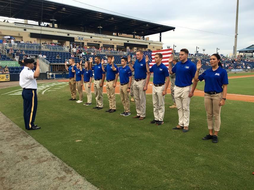 A military faculty in military uniform attire leading an outdoor oath swearing on a baseball field with a line of 10 military students holding their right hands up.