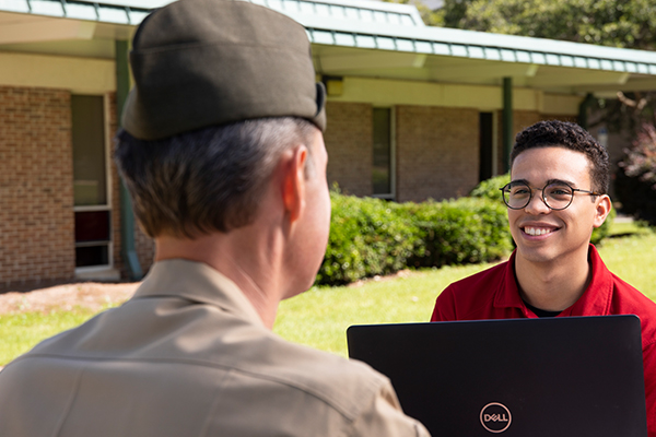 Two military students, one in military uniform and one in a polo, sitting at a table outdoors using a laptop.