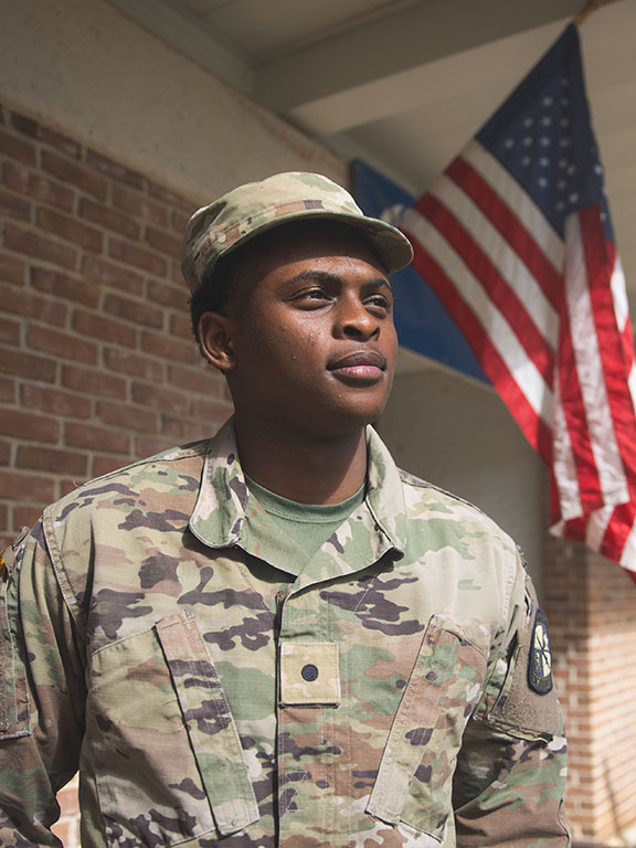 Military student in military uniform standing outdoors in front of a brick building with the U.S. flag displayed in the background.