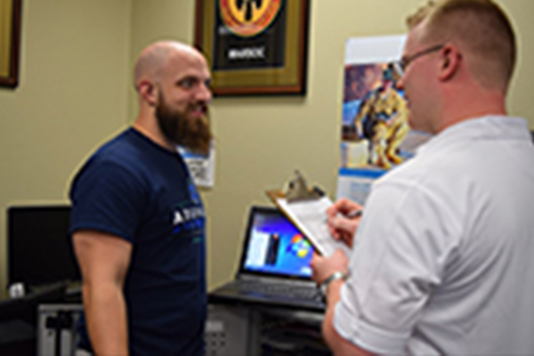 A military center staff member conversing with a military student indoors while writing on a clipboard with a laptop present in the background.