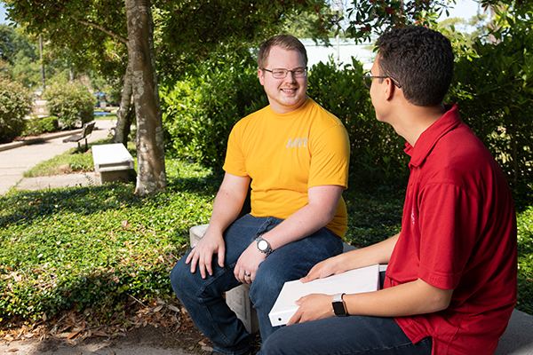 Two military students sitting at an outdoor bench conversing with various greenery around them.
