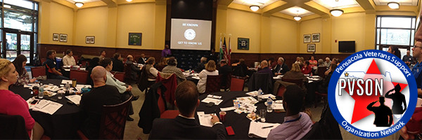 A conference room filled with attendees seated at round tables, listening to a speaker at the front, with a projector screen displaying information. The image displays a circular graphic with Pensacola Veterans Support Organization Network text on the outside and silhouettes of two military members saluting and Florida graphic with a star at Pensacola on the inside.