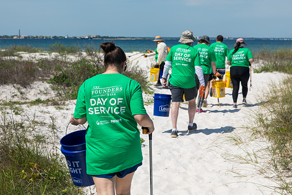 6 people walking towards the water at a beach while holding buckets and trash pickers.
