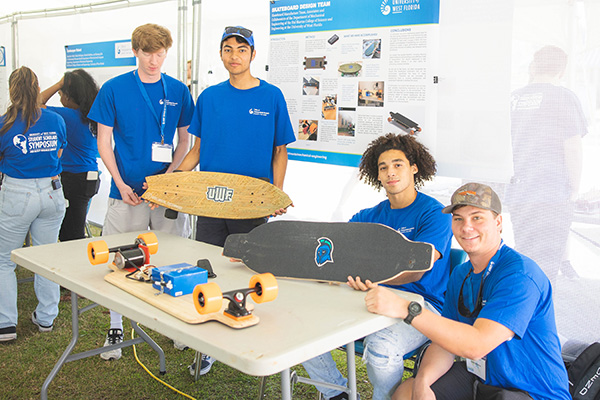 Four students presenting three battery-powered skateboards at a table during a research symposium.
