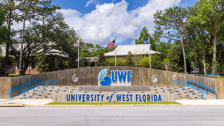 University of West Florida campus entrance monument in front of three flagpoles displaying a UWF flag, a USA flag, and a State of Florida flag from left to right.