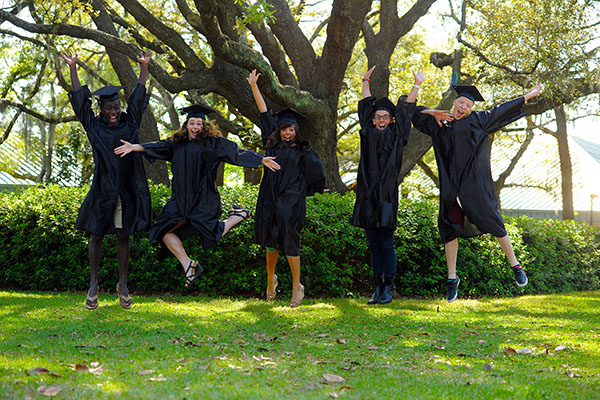 Five students in graduation regalia jumping in celebration.