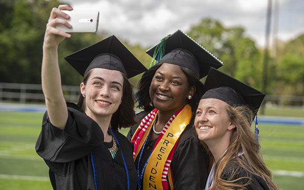 Three UWF students smile for a group phone selfie while wearing caps and gowns.