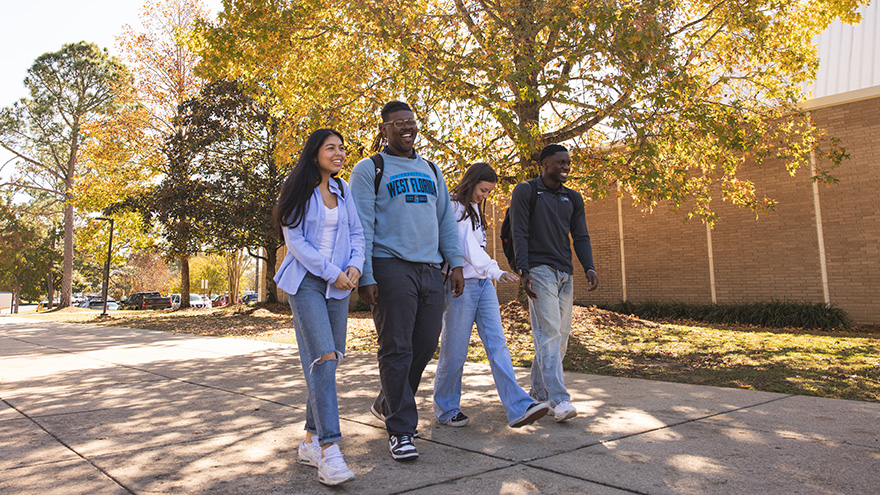 Four UWF students walking outdoors on a sidewalk with fall seasonal trees in the background.