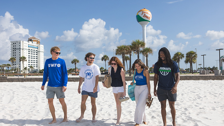 Five students walking in the sand at the beach in UWF branded attire with a condo building, volleyball nets, palm trees, and the Pensacola Beach water tower in the background.
