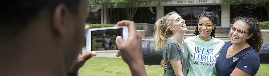 A student taking a cellphone photo of three other students in front of the cannon on Cannon Green at the UWF Pensacola campus.