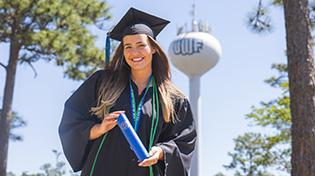 UWF graduate in cap and gown attire holding a diploma tube in front of the UWF water tower.