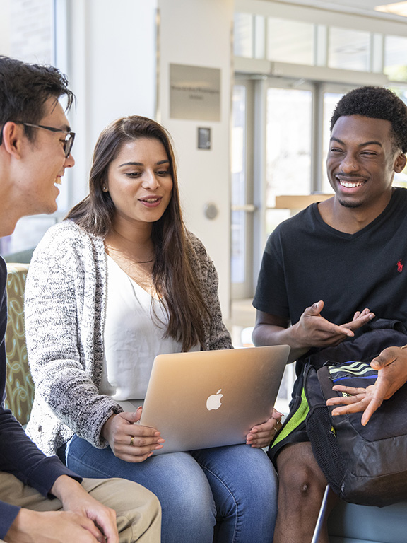 Three students conversing while sitting at the couches in Building 76A on the UWF Pensacola campus. One student is using a laptop.