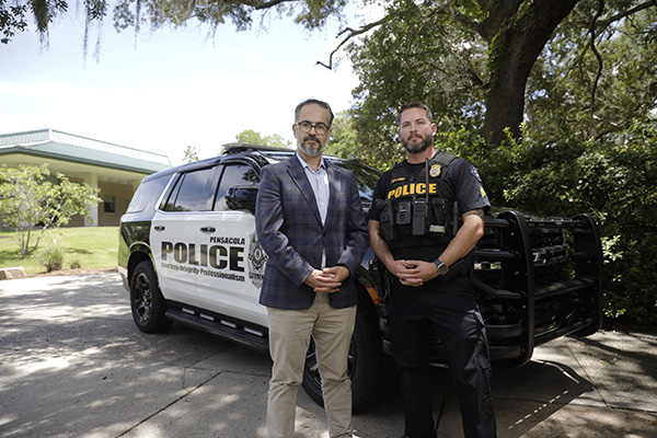 A professor in professional attire and a police officer in uniform standing outdoors in front of a police SUV.