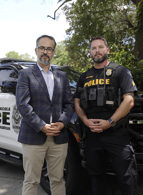 A professor in professional attire and a police officer in uniform standing outdoors in front of a police SUV.