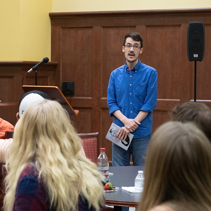 Student holding a book while speaking to a seated crowd.
