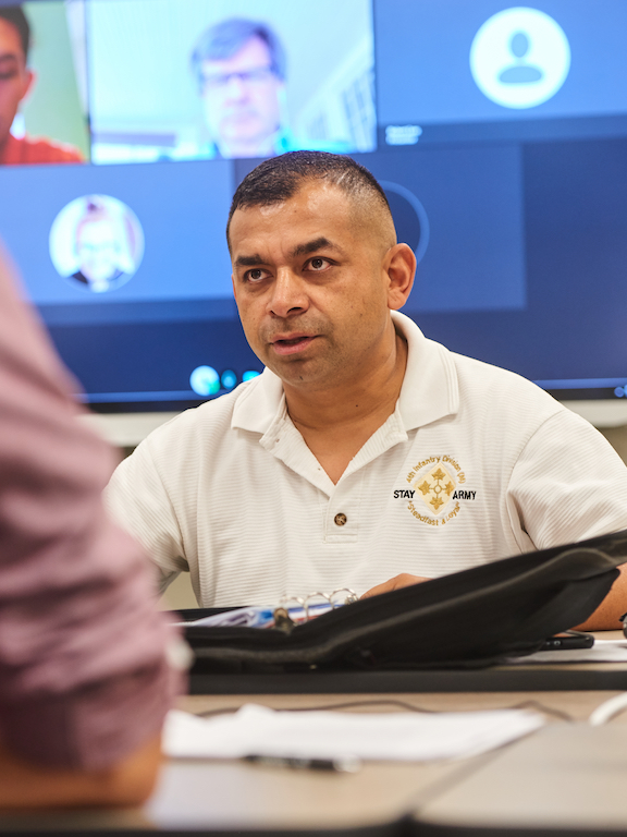 Meeting between two people at a table with a screen of virtual meeting attendees in the background.