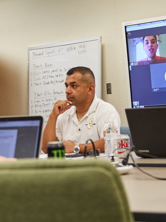 Meeting between four people at a table with a screen of virtual meeting attendees in the background.