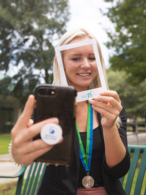 Social media professional taking a selfie outdoors with a smartphone while holding a small #UWF24 cutout frame in front of their face.