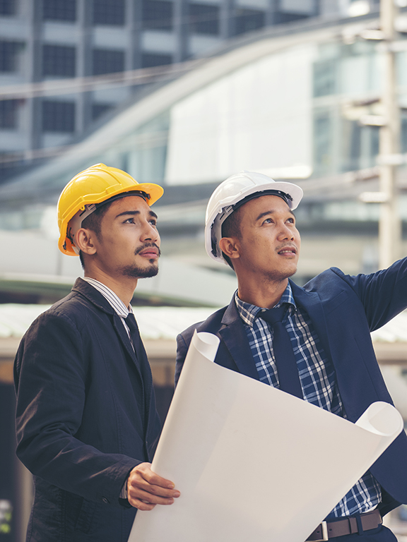 Two civil engineers in professional attire wearing hardhats discussing blueprints.