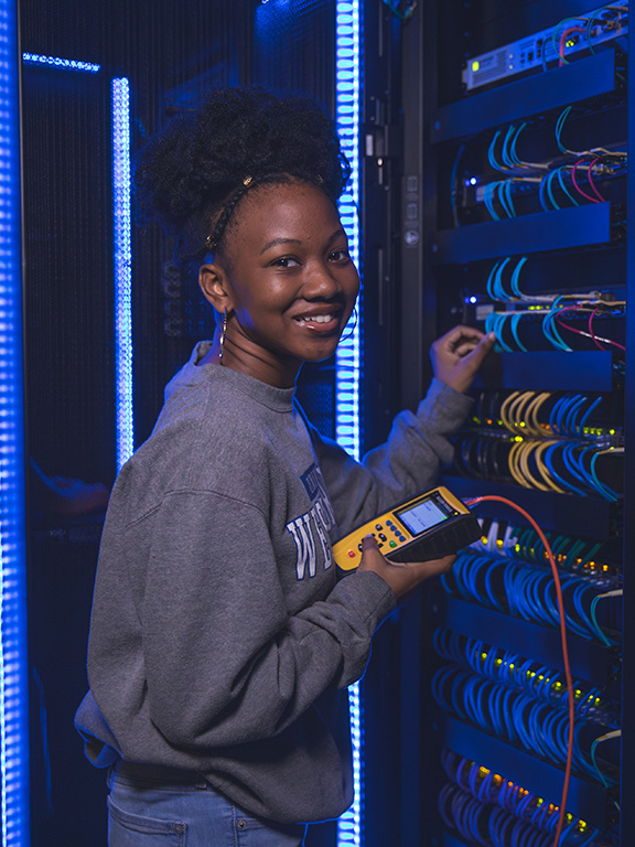 A student smiling at camera while working on cybersecurity related computer equipment.