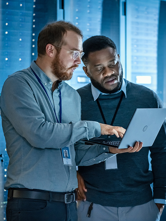 Two information technology professionals standing in front of a web servers conversing together while one is holding a laptop.