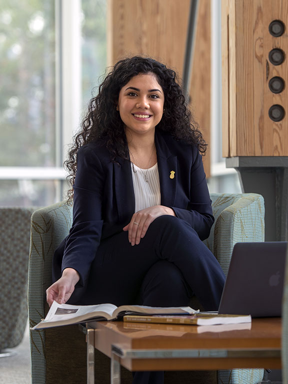 A student in professional attire smiling at camera while sitting in a chair and flipping a page of a textbook that is placed on a small table in front of the student.