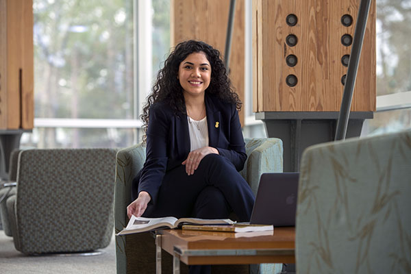 A student in professional attire smiling at camera while sitting in a chair and flipping a page of a textbook that is placed on a small table in front of the student.