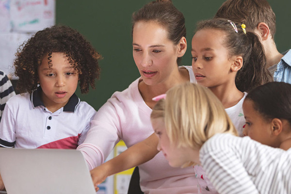 Five young students crowded around a teacher to look at a laptop screen.
