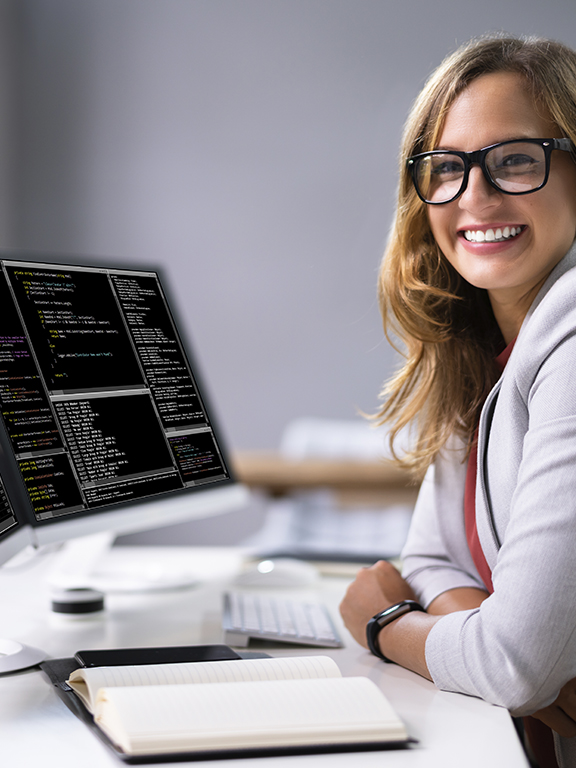 Programmer smiling in front of two computer monitors that display various software code.