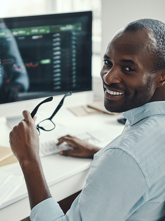 Data analyst professional smiling holding a pair of glasses while working on a computer with data displaying on screen.