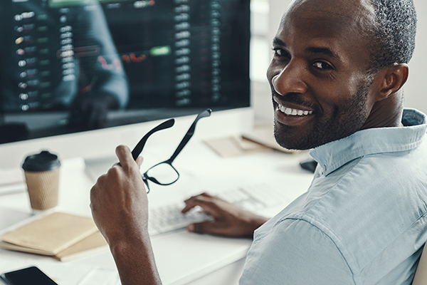 Data analyst professional smiling holding a pair of glasses while working on a computer with data displaying on screen.