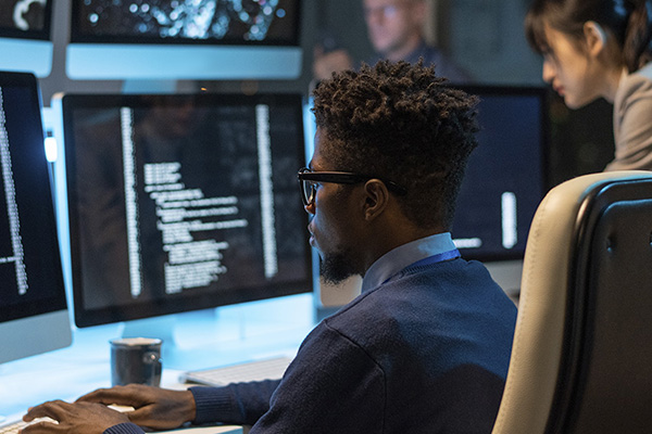 Information security manager sitting in front of computer monitors that display software code.