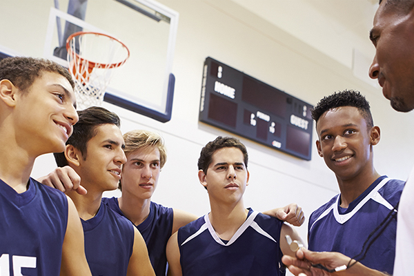 Close up of a high school basketball team talking to a coach with a basketball hoop and scoreboard in the background.