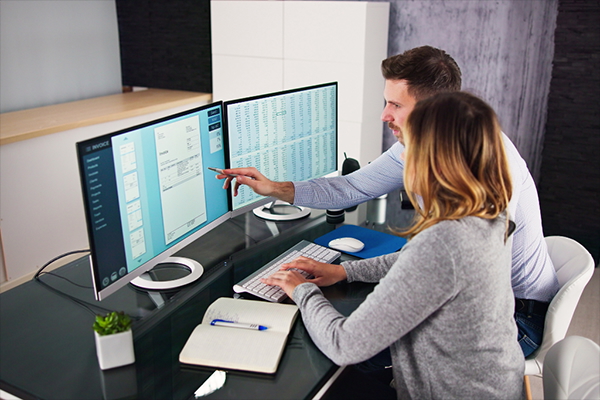 Two financial planners using a computer with two monitors to review an invoice and a financial spreadsheet.